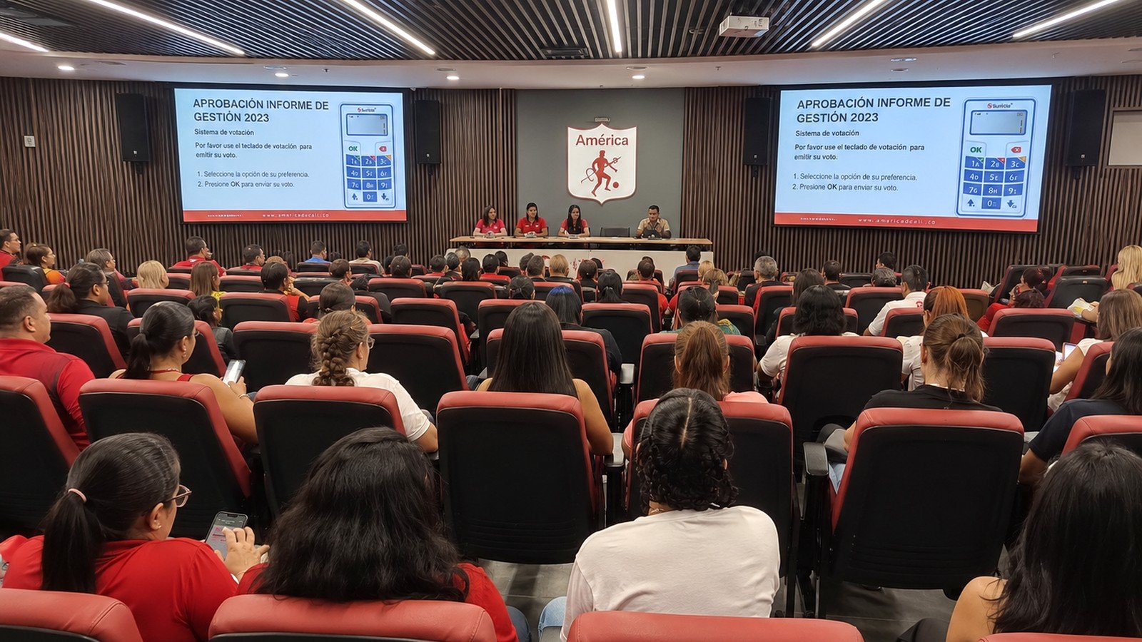 Sala de la Asamblea General Ordinaria 2026 de América de Cali con accionistas, doble pantalla y votación con controles físicos.