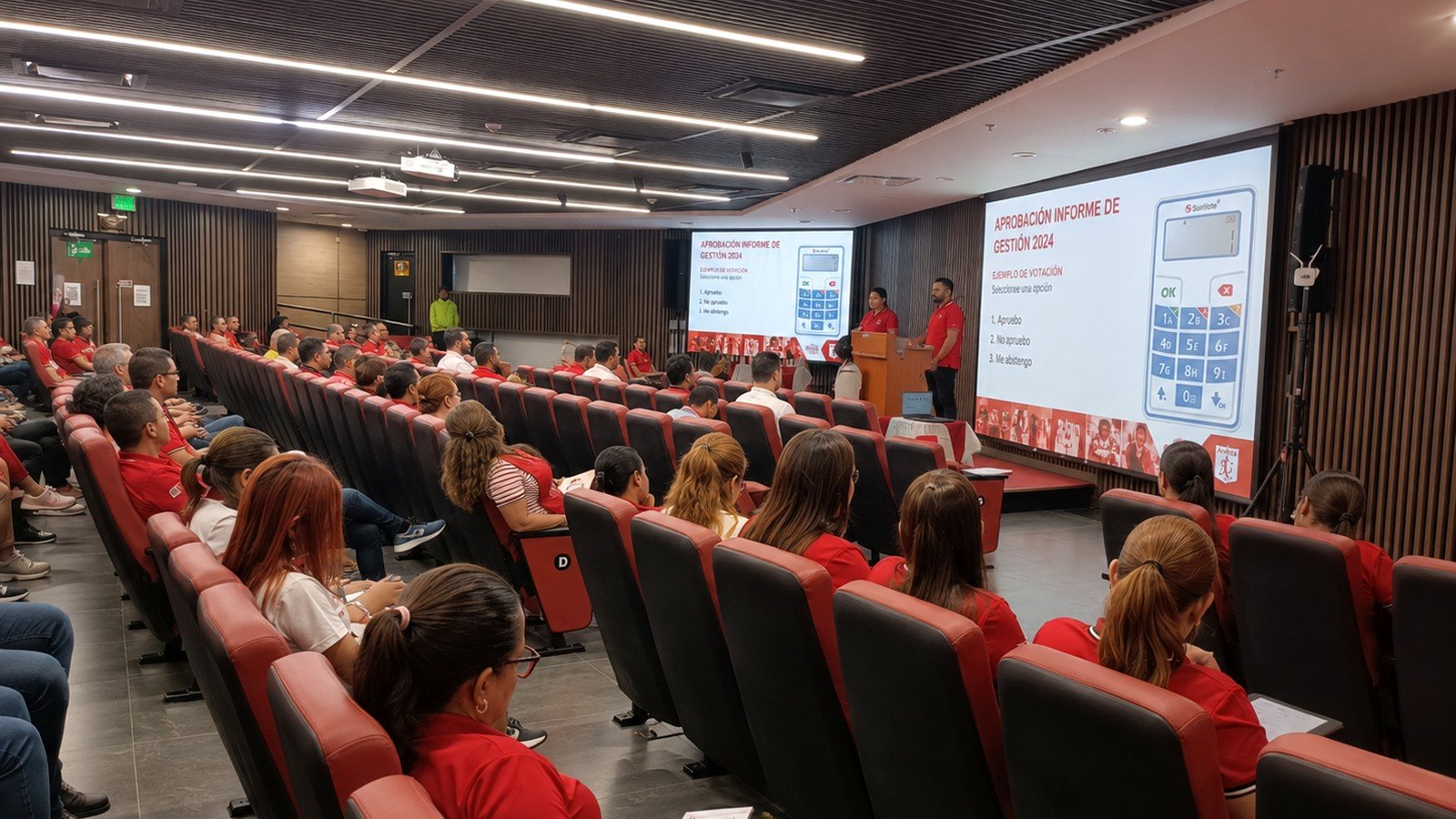 Asamblea de América de Cali con asistentes e instrucciones de votación electrónica proyectadas en pantalla.