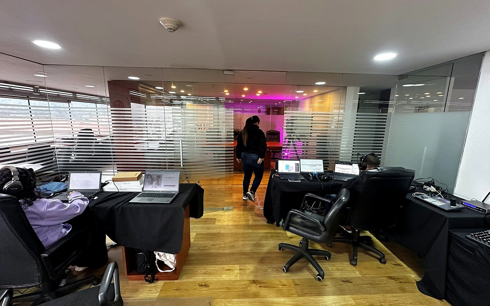 Montaje de estudio para asamblea Colpeunidos con mesas técnicas, laptops, cámara PTZ y pantalla de apoyo.